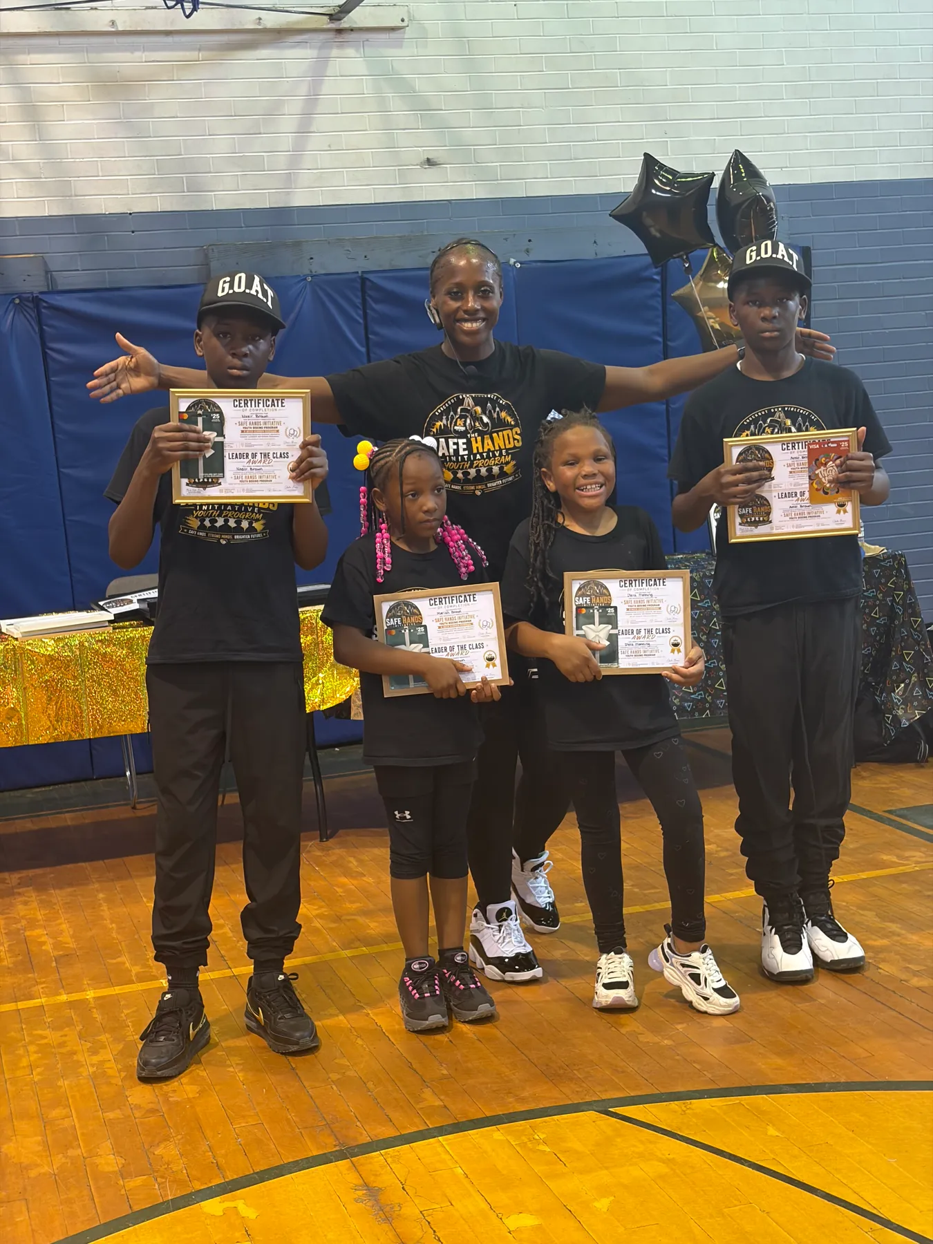 Fisher with four children holding framed Safe Hands Initiative certificates in a recreation center gym.